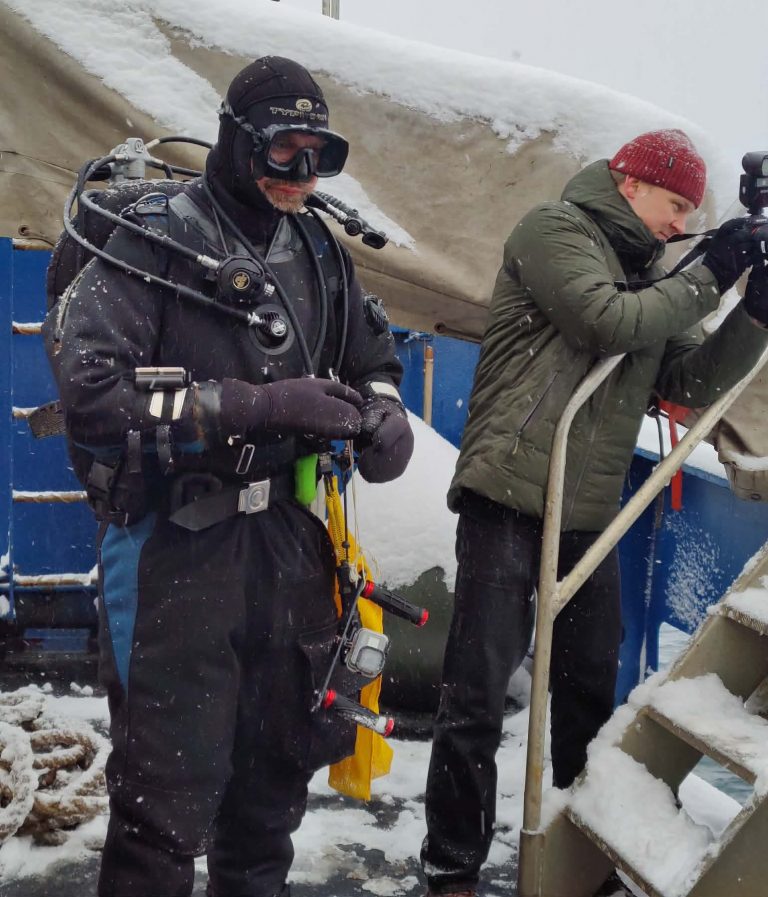 Polar diver in a drysuit preparing equipment on deck in snowy conditions with team support in Greenland.