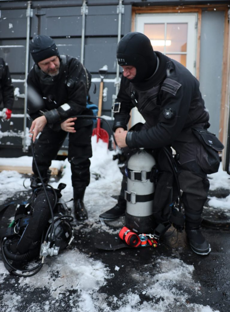 Two polar divers in drysuits preparing dive gear on ice in winter conditions in Greenland.