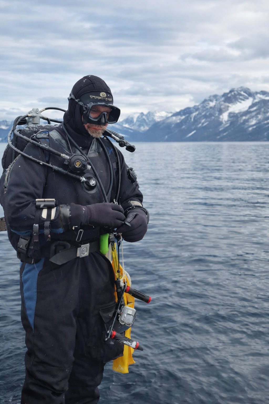 Cold-water diver in drysuit preparing equipment on deck with Arctic water and mountains in the background.