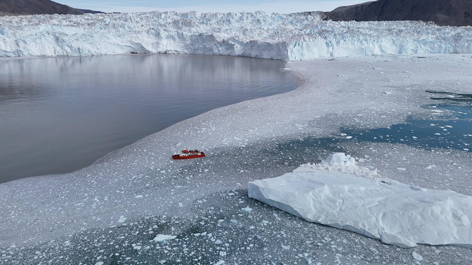 Small expedition boat navigating sea ice near a massive glacier wall in Greenland