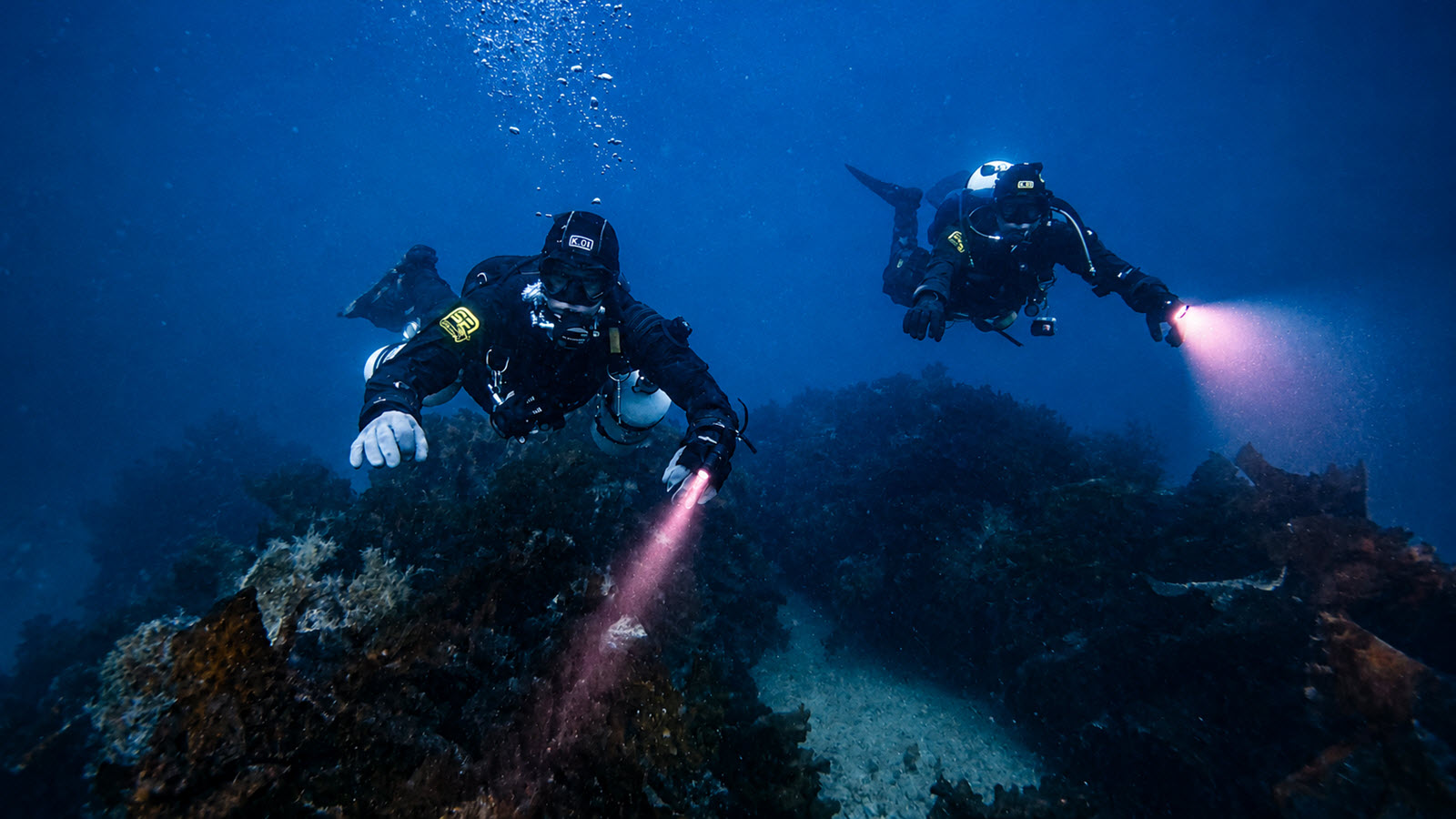 Two divers with lights exploring a rocky reef in search of a shipwreck