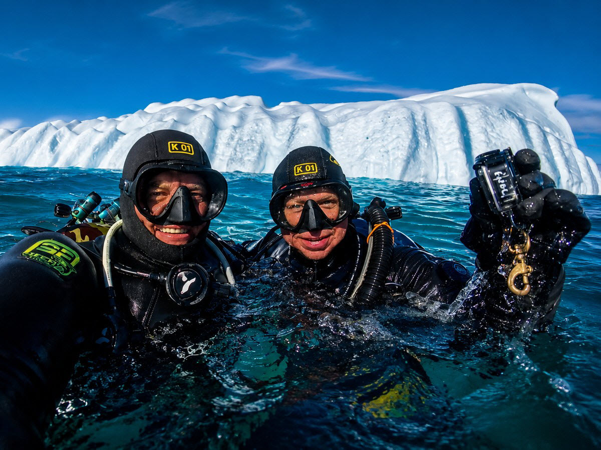Two divers in drysuits surfacing beside an iceberg in Greenland