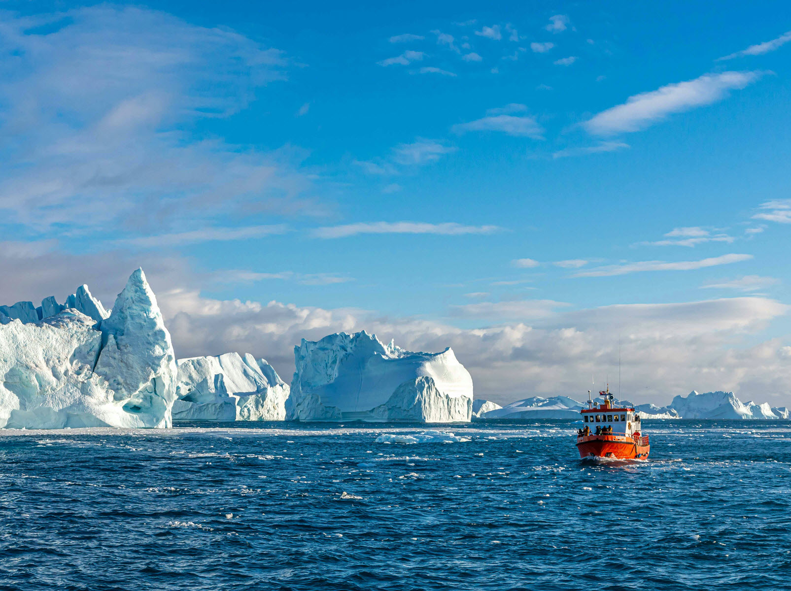 A small red boat passing towering icebergs in Greenland