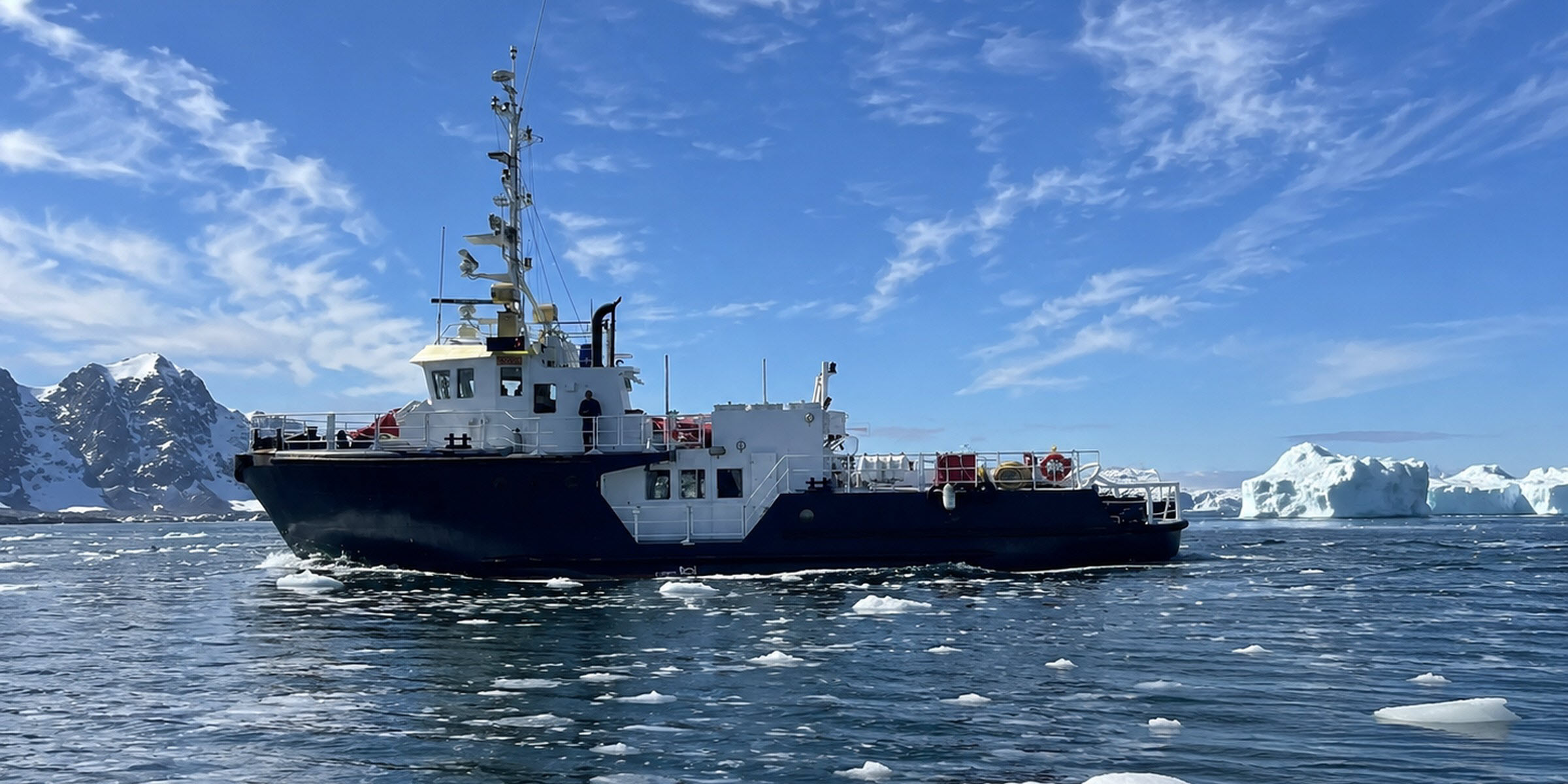 M/V Sisak underway through pack ice past an iceberg and snow-capped peaks in Greenland