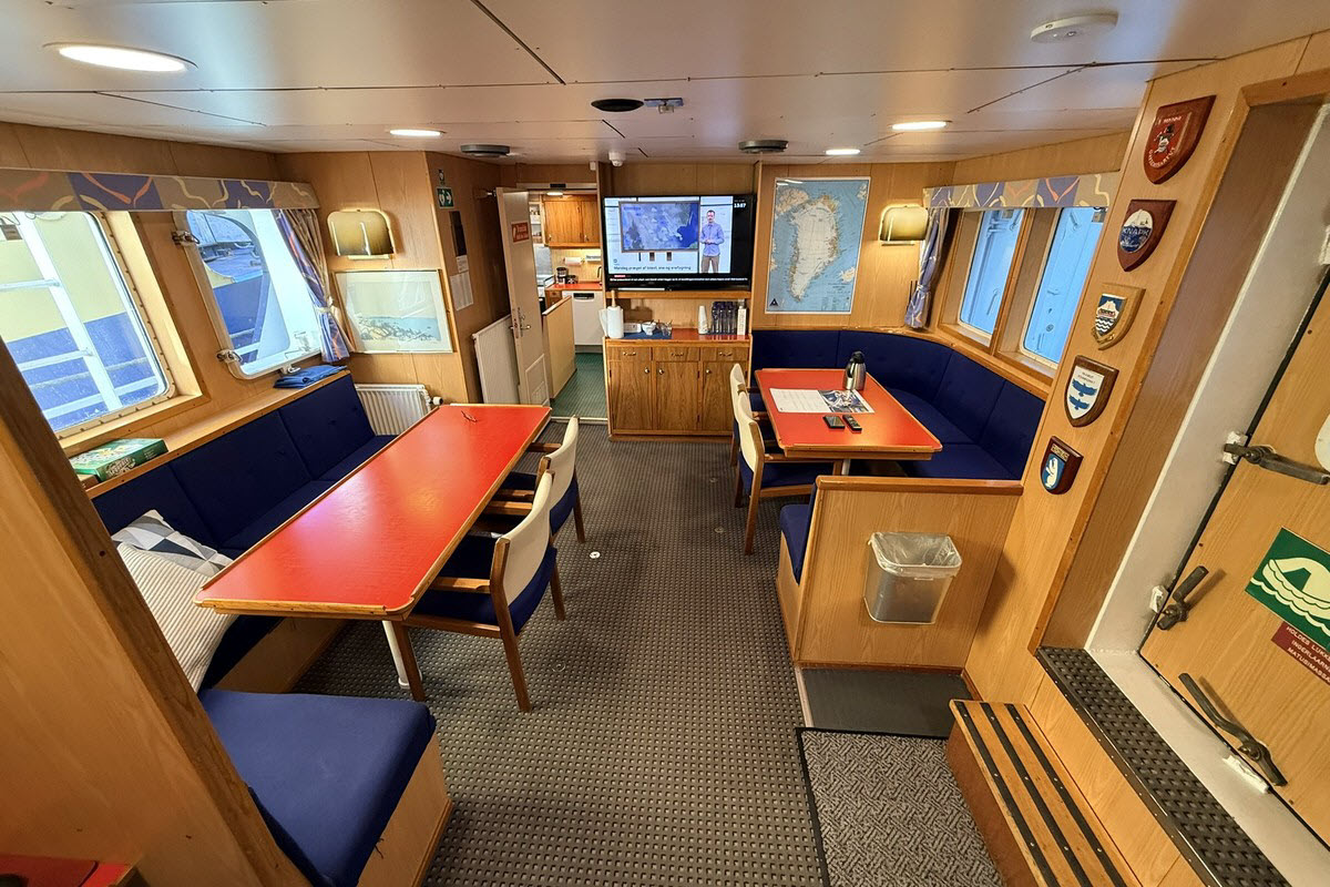 The mess and saloon aboard M/V Sisak with dining tables, banquette seating, and a Greenland chart on the wall