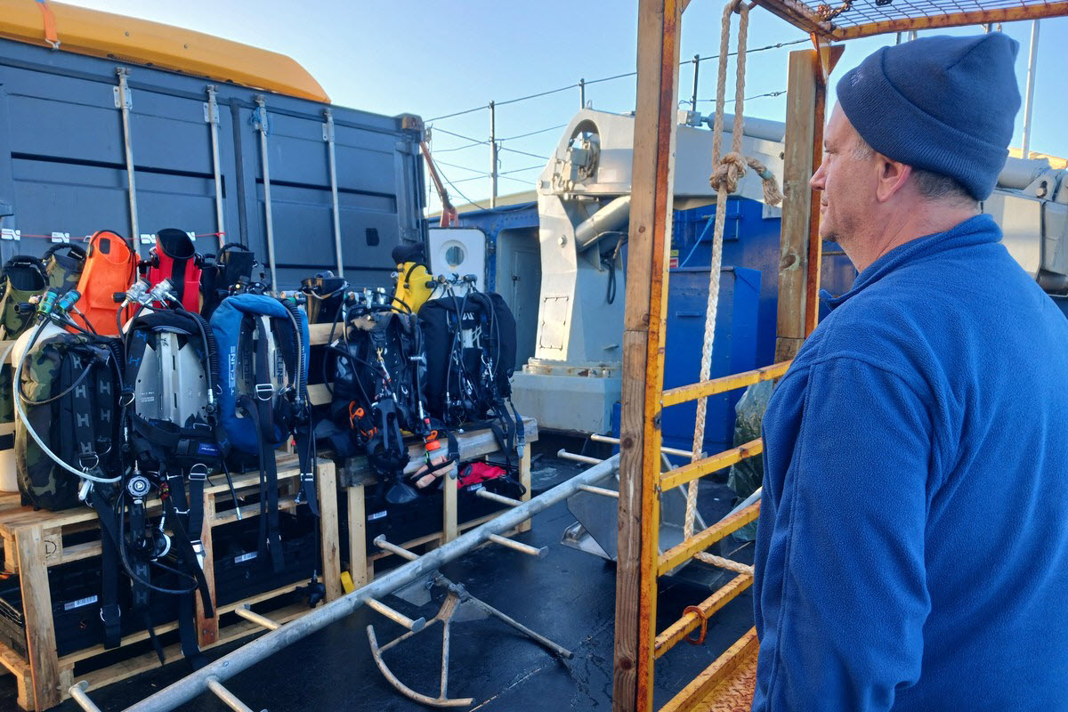 Rows of scuba rigs and drysuits staged on deck aboard M/S Tulu as a diver looks on