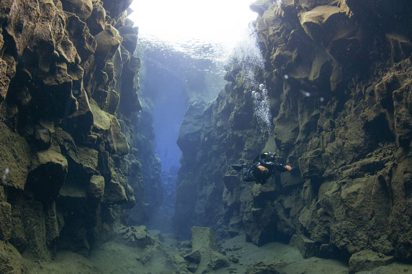 A diver drifting through the Silfra tectonic fissure between basalt walls in Iceland, light pouring down from the surface