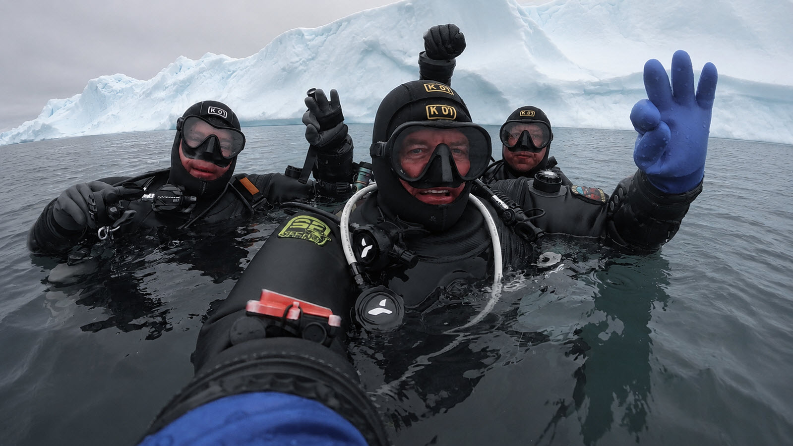 Three divers surfacing together beside an iceberg, giving the OK signal
