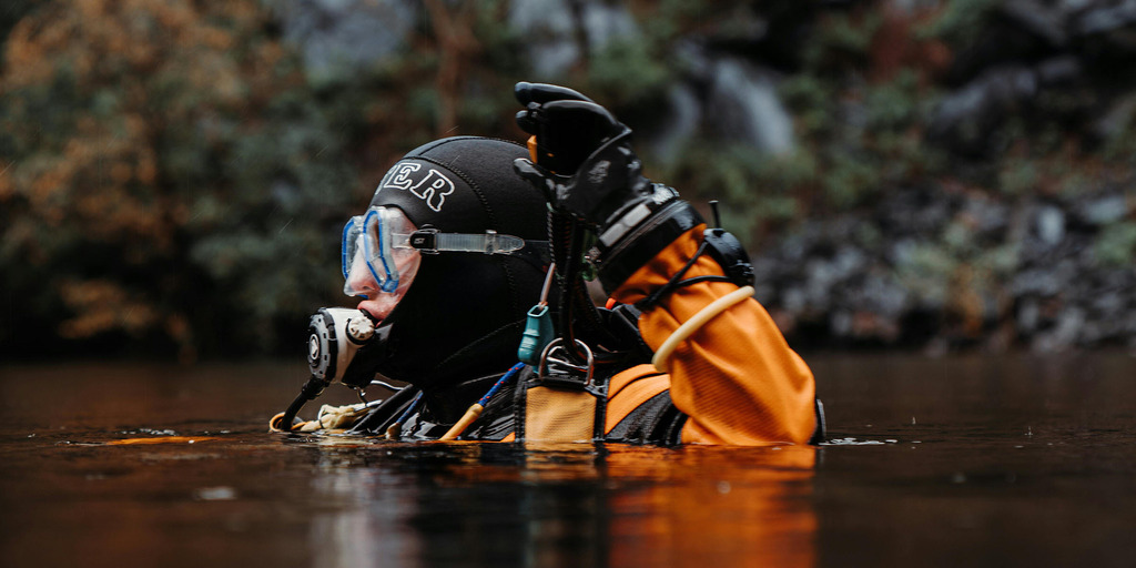 A drysuit diver at the surface of a dark river beneath a waterfall