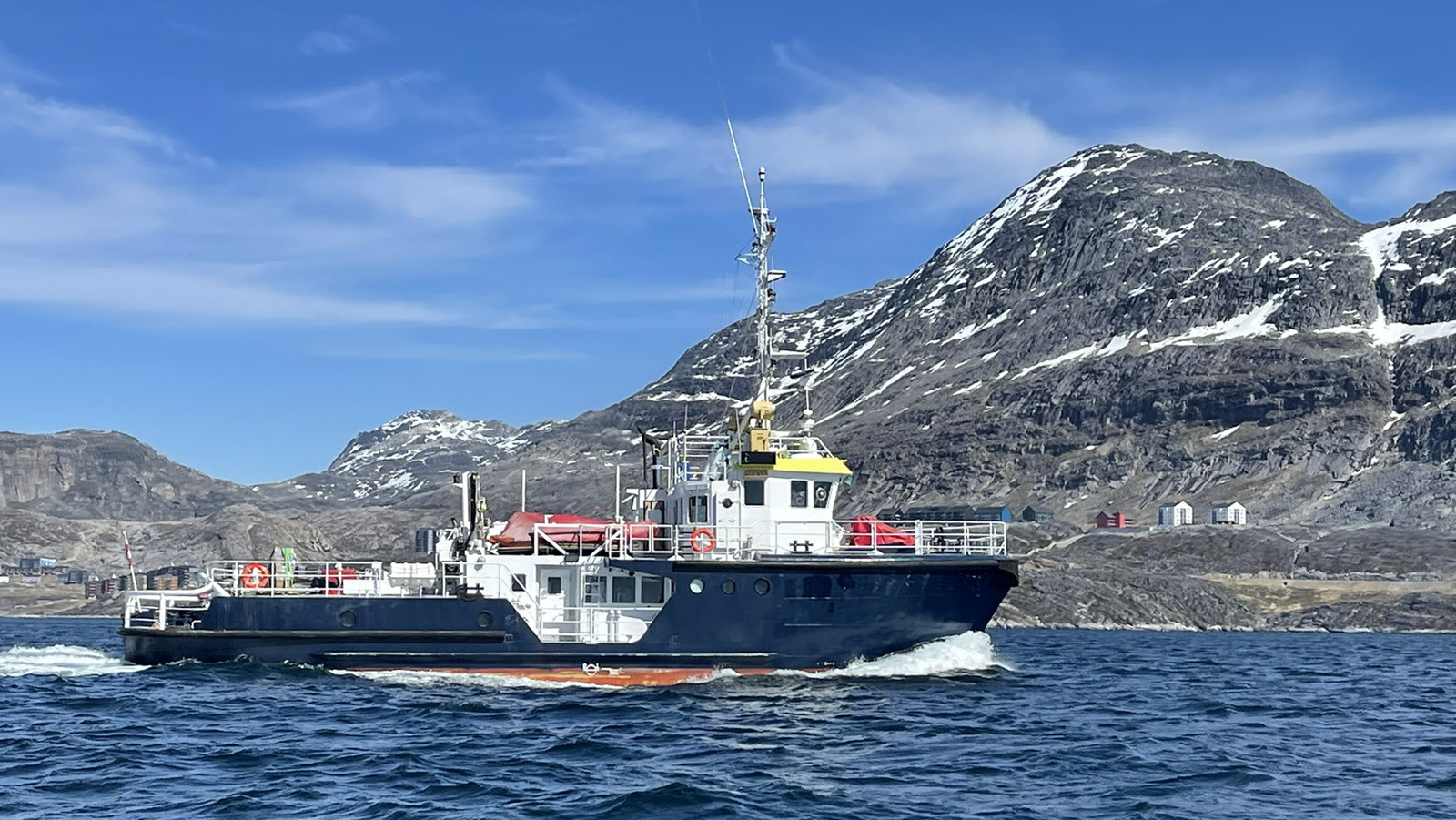 M/V Sisak underway in a Greenland fjord below snow-streaked mountains, with a coastal settlement on the shoreline
