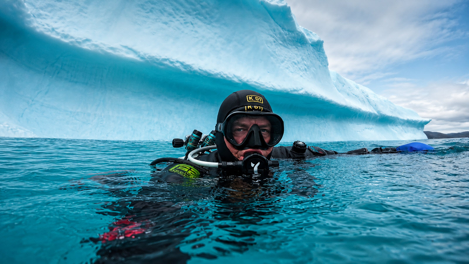 A drysuit diver at the surface beside a large turquoise iceberg in Greenland